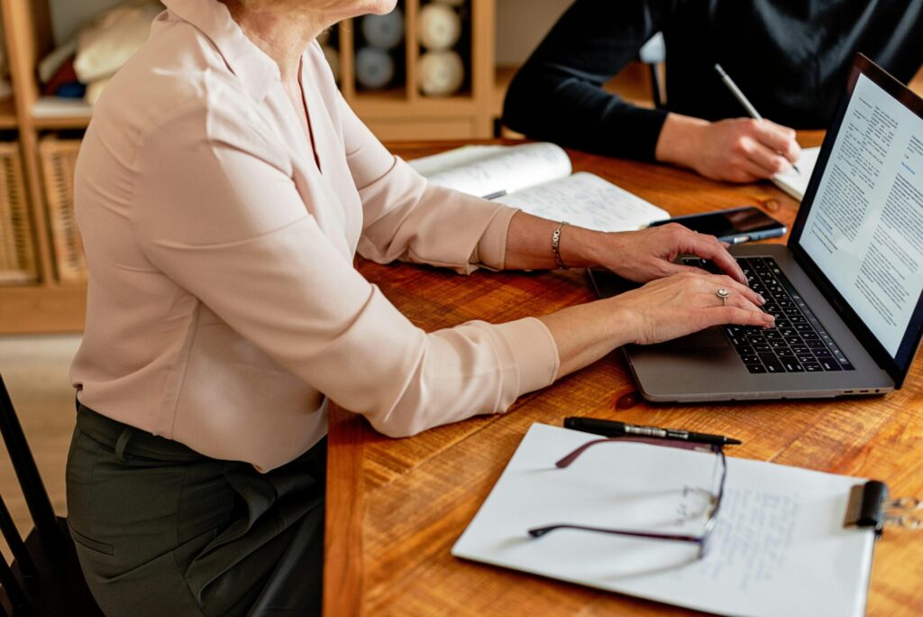 Two professionals at a desk, using a laptop and taking notes in a collaborative work setting.