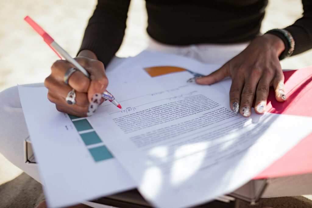 A close-up shows a person editing papers outdoors using a red pen, hinting at a creative review process.
