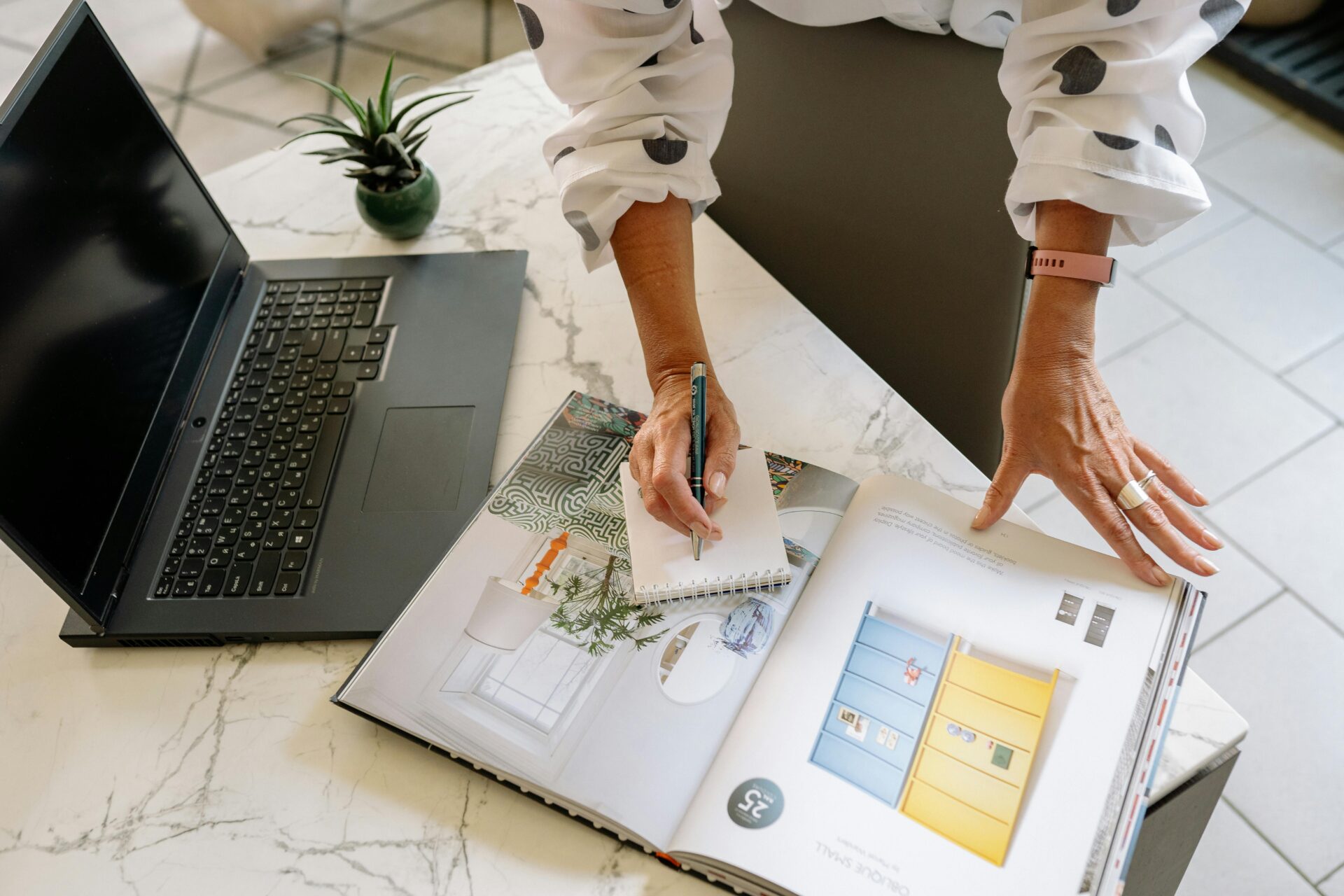 Businesswoman taking notes from a magazine on a marble surface with a laptop nearby.