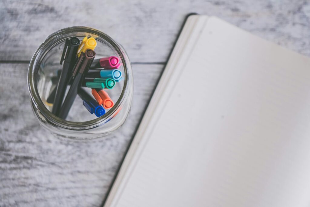 Top view of a glass jar filled with colorful pens next to an open notebook on a wooden desk.